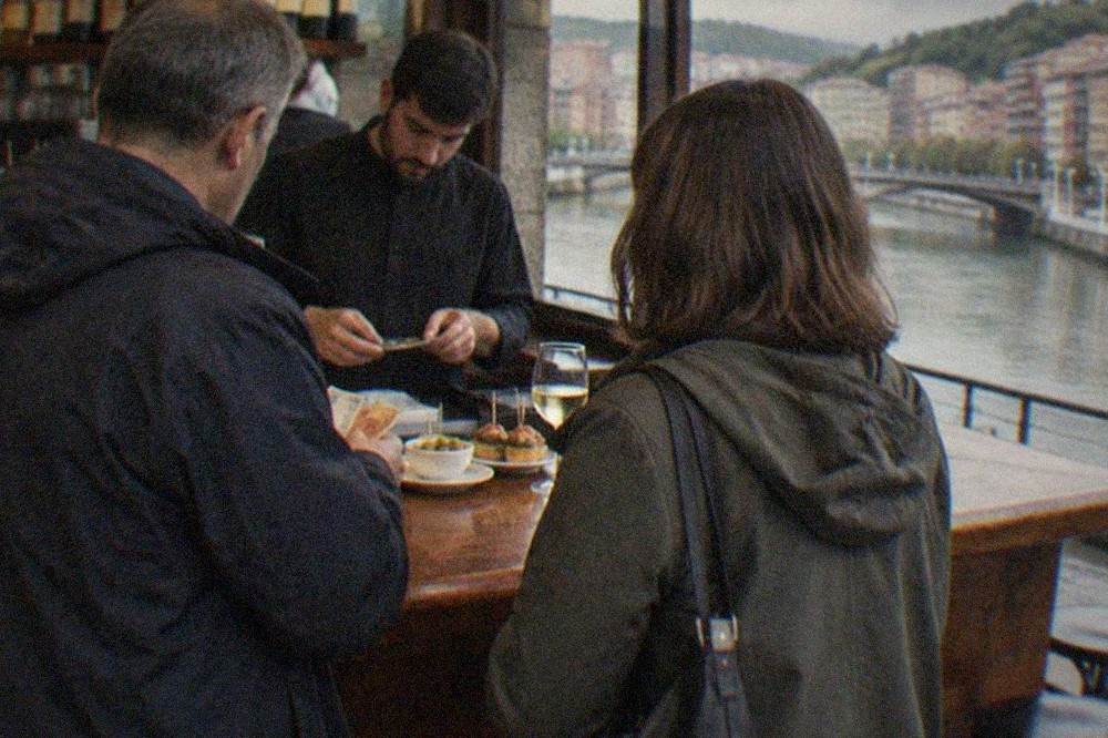 Two people standing at a Bilbao pintxos bar counter paying their bill with cash while the barman makes change