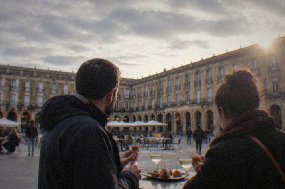 Two people standing at a Plaza Nueva bar in Bilbao eating skewered pintxos with small glasses of txakoli