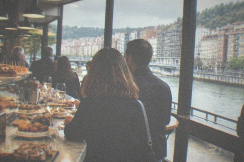 A busy pintxos bar counter in Bilbao with plates of small bites, glasses of txakoli wine and standing customers