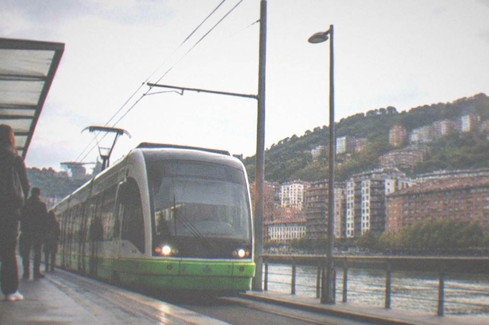 A Bilbao tram passing modern apartment buildings along the estuary with passengers waiting at a platform