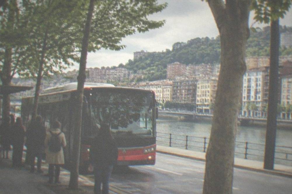 A Bilbao city bus at a stop on a tree-lined street, with passengers waiting at the kerb