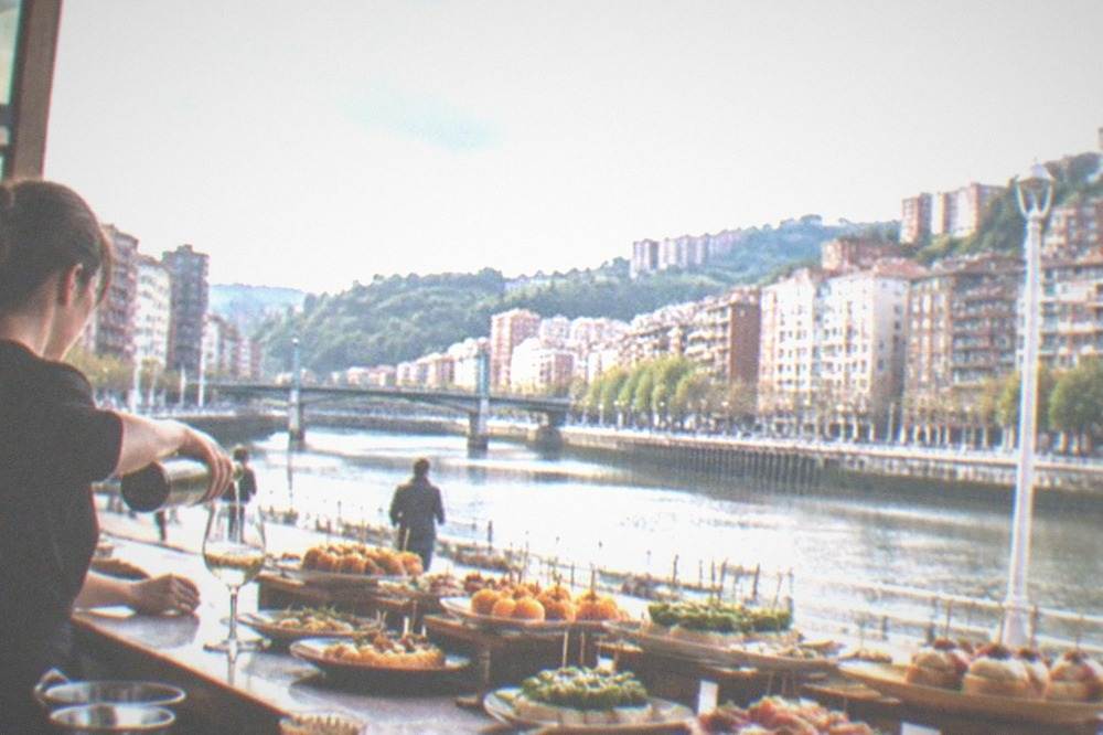A pintxos bar counter in Bilbao loaded with small plates on skewers, with a bartender pouring txakoli wine