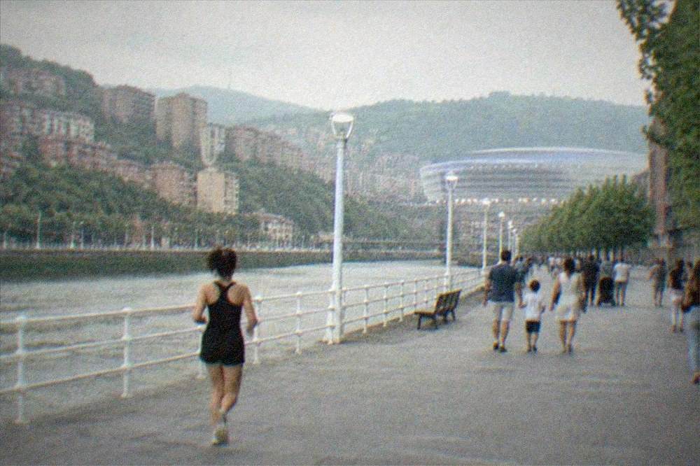 Families walking along a wide boulevard in Bilbao on a summer evening, with the stadium's lights visible ahead