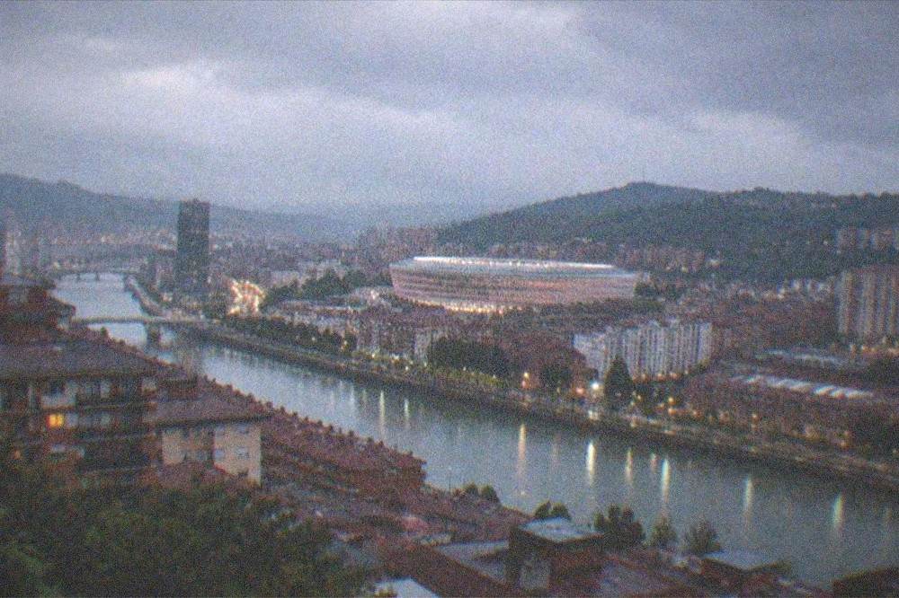 A twilight view over western Bilbao rooftops with the illuminated stadium and distant city lights