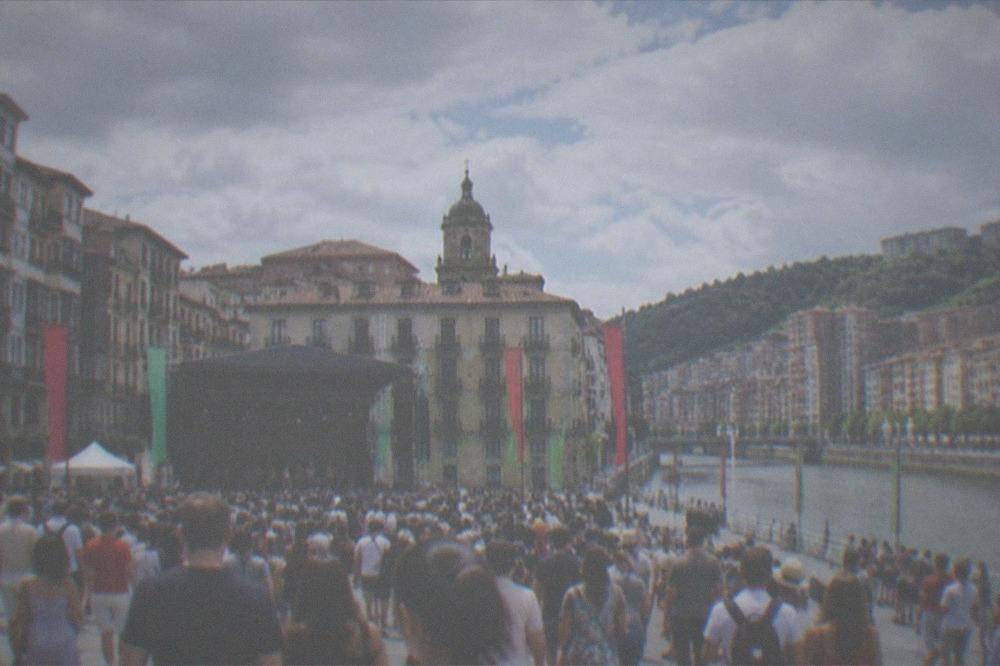 Crowds filling a Bilbao old town square during a summer festival with flags and stage structures