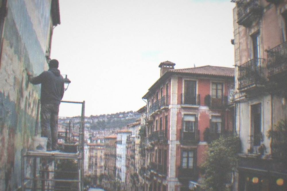 A painter working on a large mural on a brick wall while pedestrians pass on the pavement