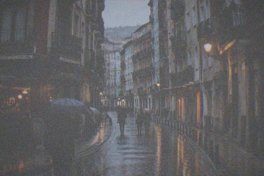 Wet cobblestone street reflecting lights in the evening after rain, with people under umbrellas