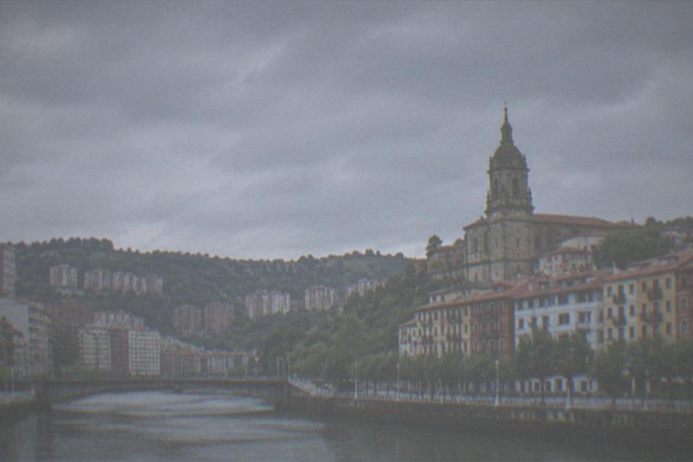 A hilltop church tower against a cloudy sky above old town rooftops in Bilbao