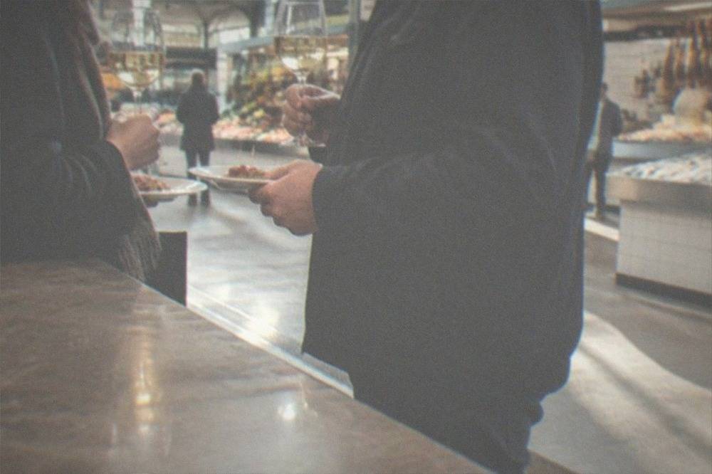 Two people standing at a bar counter holding small plates and glasses of white wine