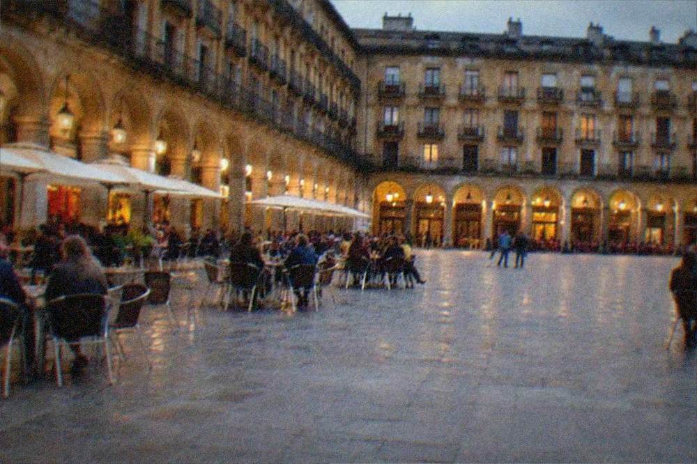 Evening view of an arcaded square with lit bar windows and people gathered at outdoor tables