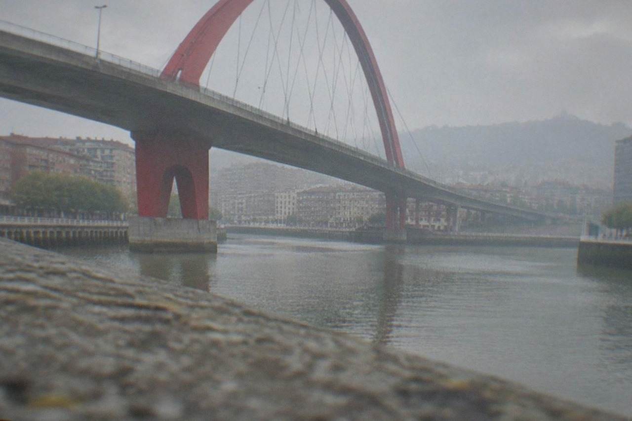 A tall red bridge with a curved arch crossing a river between buildings in a Spanish city