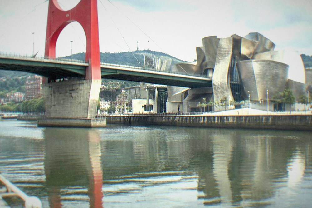 A red bridge arch reflected in a city river beside a modern metallic museum building