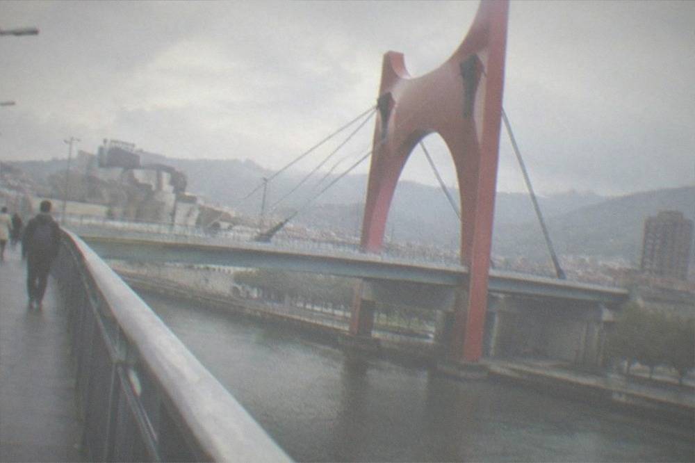Pedestrians walking on a bridge sidewalk above a city river on an overcast day