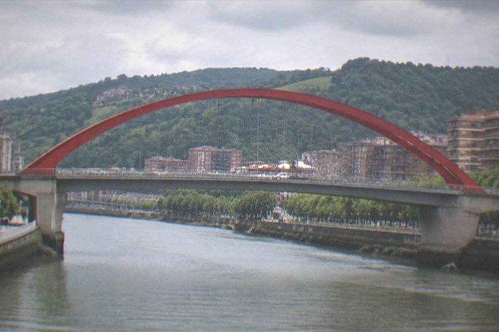 A red arched bridge over a river with green hills behind a northern Spanish city