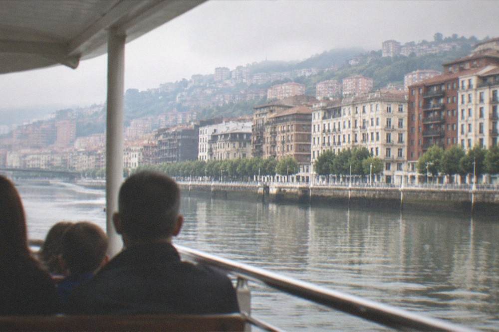 A family on a covered river boat looking out at the Bilbao riverside buildings