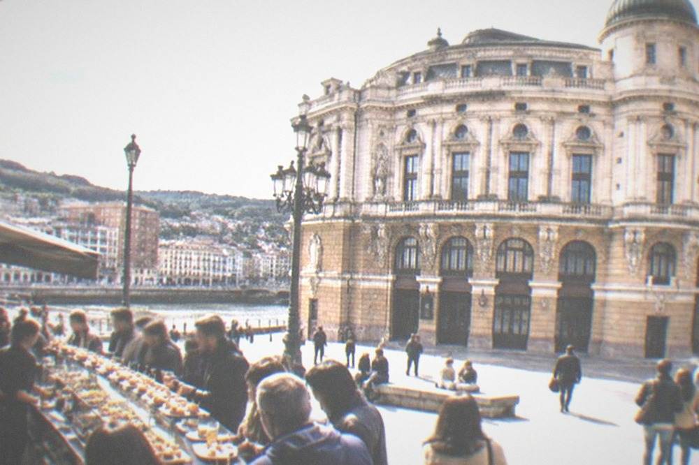 People eating pintxos at a crowded bar counter in an arcaded square near the theatre