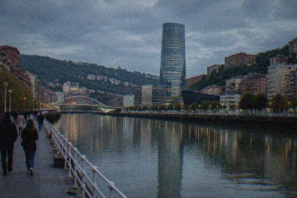 Visitors walking along the riverside promenade in Bilbao with the glass tower and modern buildings reflected in the estuary water
