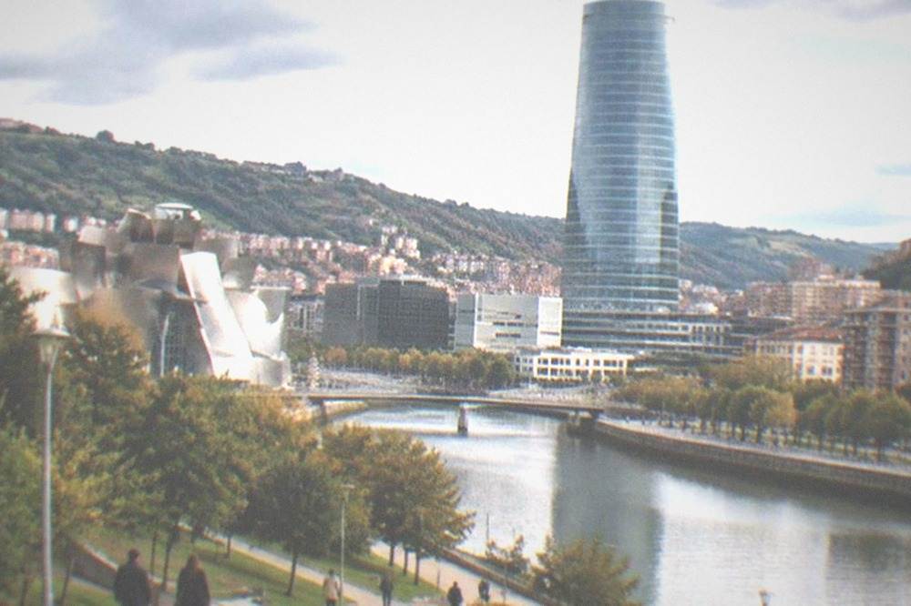 The glass tower in Bilbao under a partly cloudy Atlantic sky with green lawns and pedestrians below