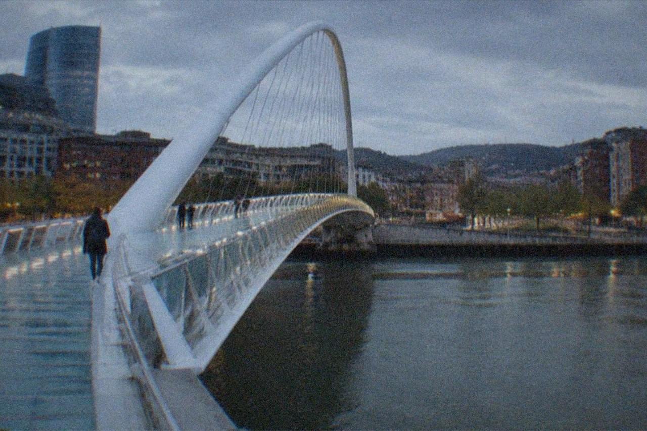 A white curved footbridge arching over a city river with modern buildings on both banks