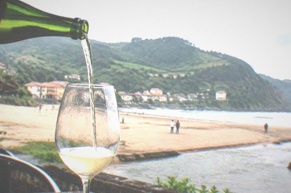 A glass of pale txakoli wine being poured high into a glass at a seaside bar