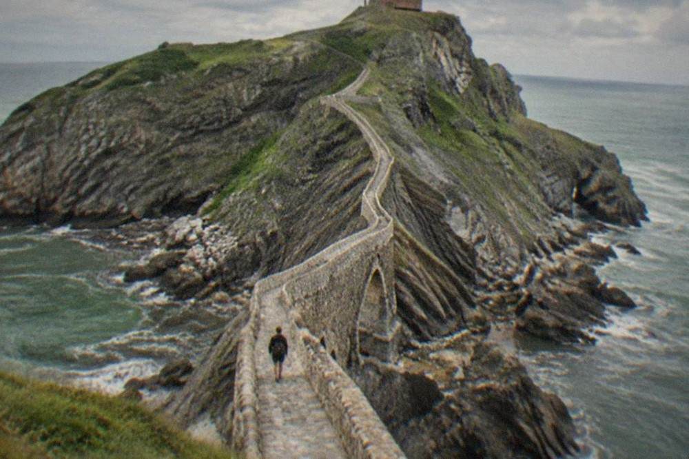 A visitor walking the narrow stone bridge toward the staircase cut into the rock at San Juan de Gaztelugatxe