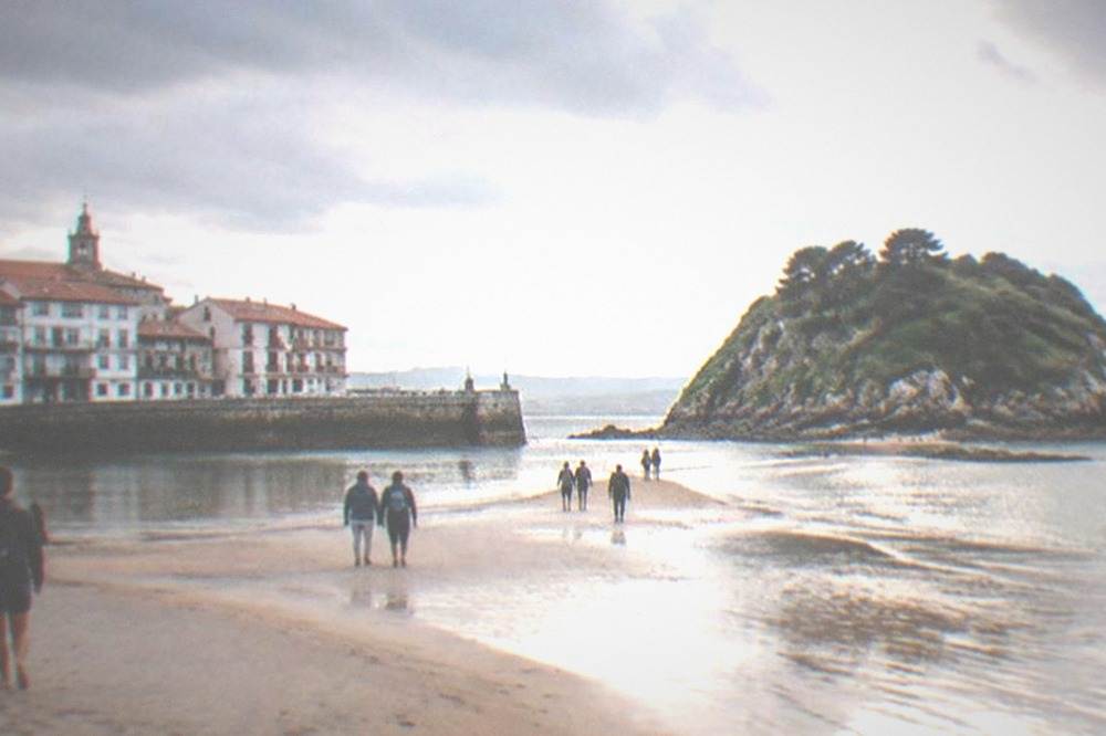 People walking across exposed wet sand toward a small green island from a town beach