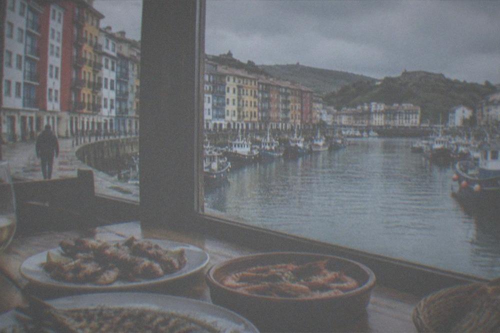 Plates of grilled fish and seafood on a wooden table beside a window overlooking a fishing port