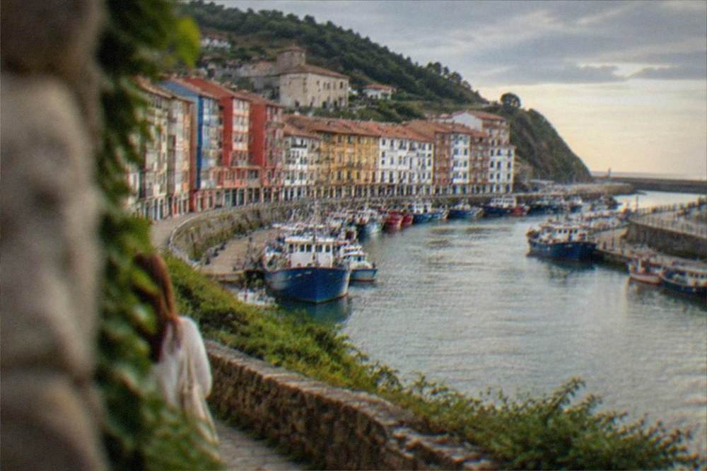 A Basque coastal town seen from a headland path under a bright summer sky with calm sea