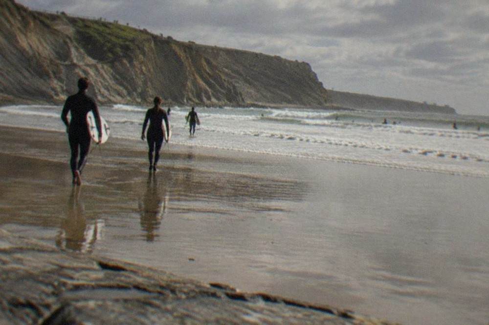 Surfers in wetsuits walking across wet sand toward breaking waves