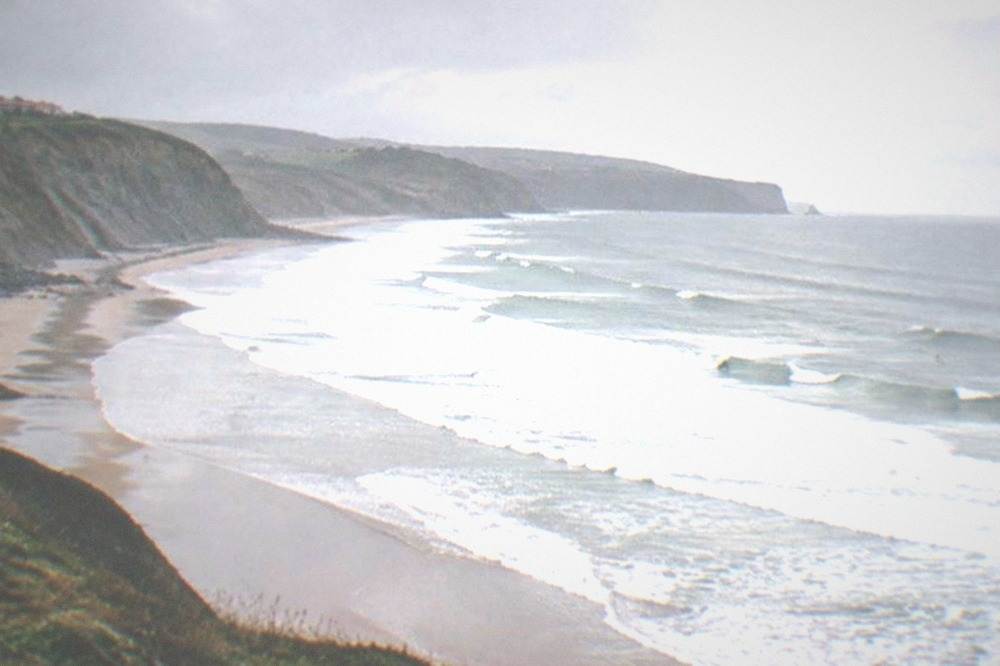 View from a cliff over a long curving beach with white water and a grey Atlantic sky