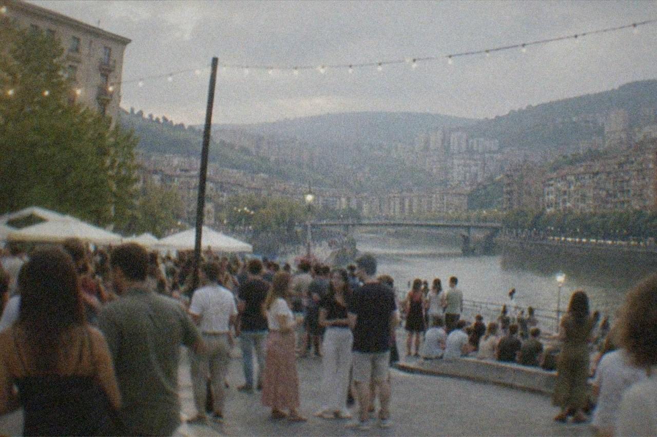 Crowds gathered in a Bilbao square at dusk during a summer festival with lights strung above the street