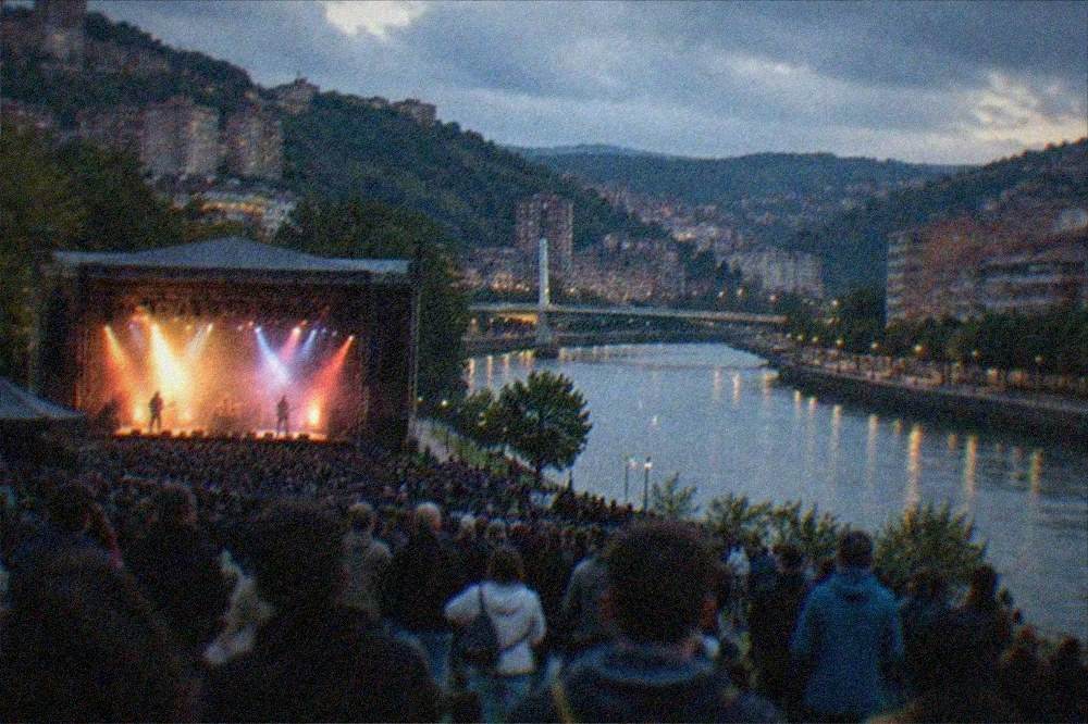 An outdoor concert crowd facing a lit stage in a riverside park at night