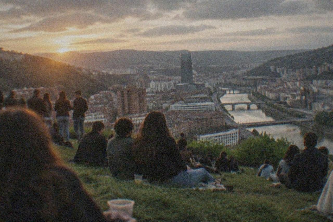 Crowd of festivalgoers on a green hillside above Bilbao at dusk with the city skyline below