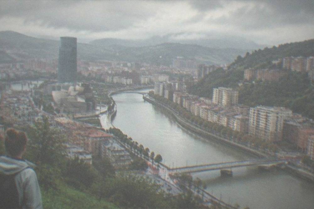 Person with a small backpack wearing a light rain jacket on a green hillside under a partly cloudy sky