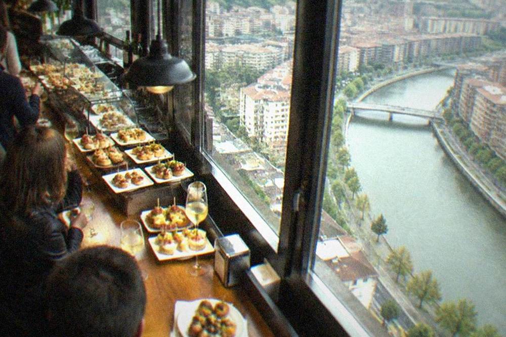 A crowded pintxos bar counter with small skewered bites and glasses of txakoli
