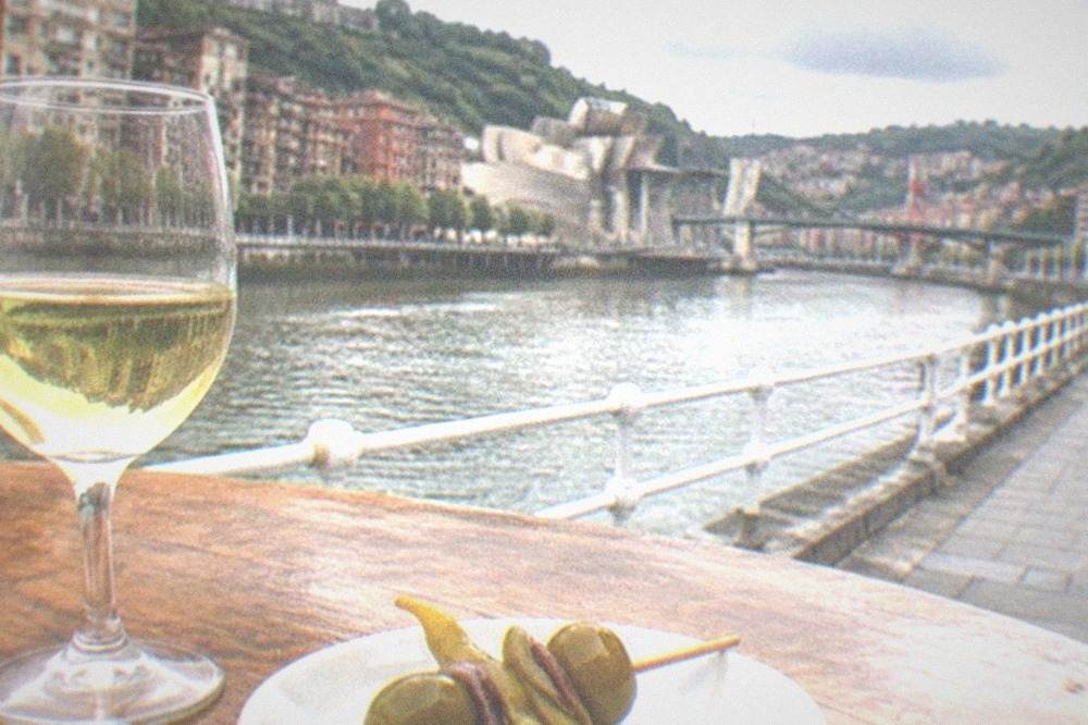 A close-up of a gilda skewer and a glass of pale txakoli on a wooden bar top