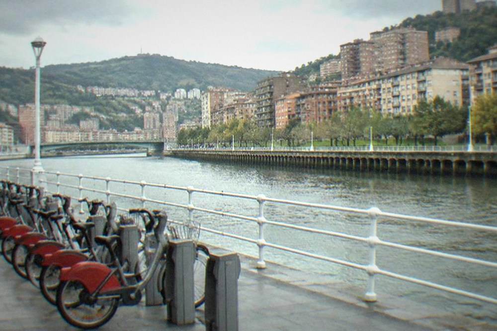 A row of rental city bikes parked on a wide riverside promenade in Bilbao on a dry overcast day