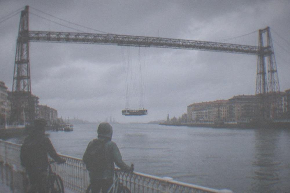 Two cyclists on a riverside path looking up at the Vizcaya transporter bridge spanning the water at Portugalete