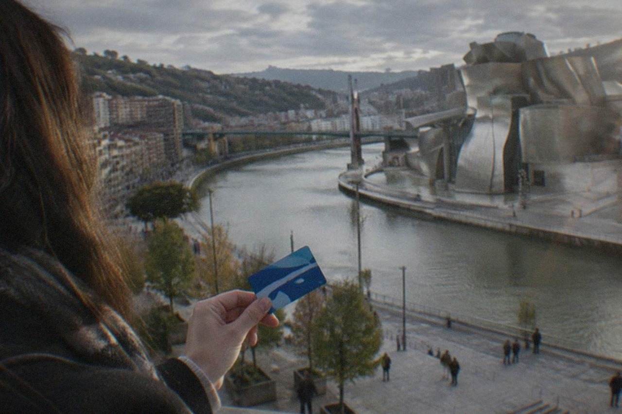 A tourist holding a city transport card while standing in a Bilbao plaza with the Guggenheim Museum's titanium curves in the background