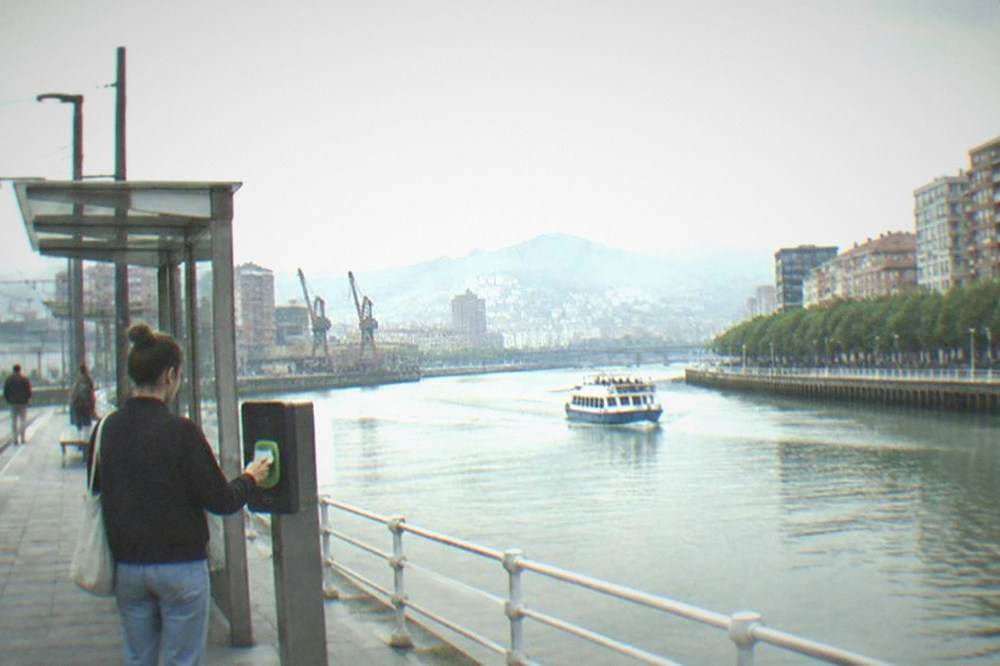A visitor tapping a transport card on a validator at a Bilbao tram stop beside the Nervion river