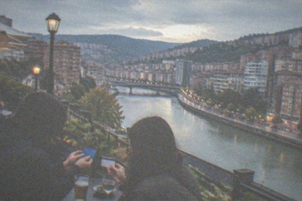 Two travellers comparing paper transport cards at an outdoor table in a Bilbao square lined with pintxos bars