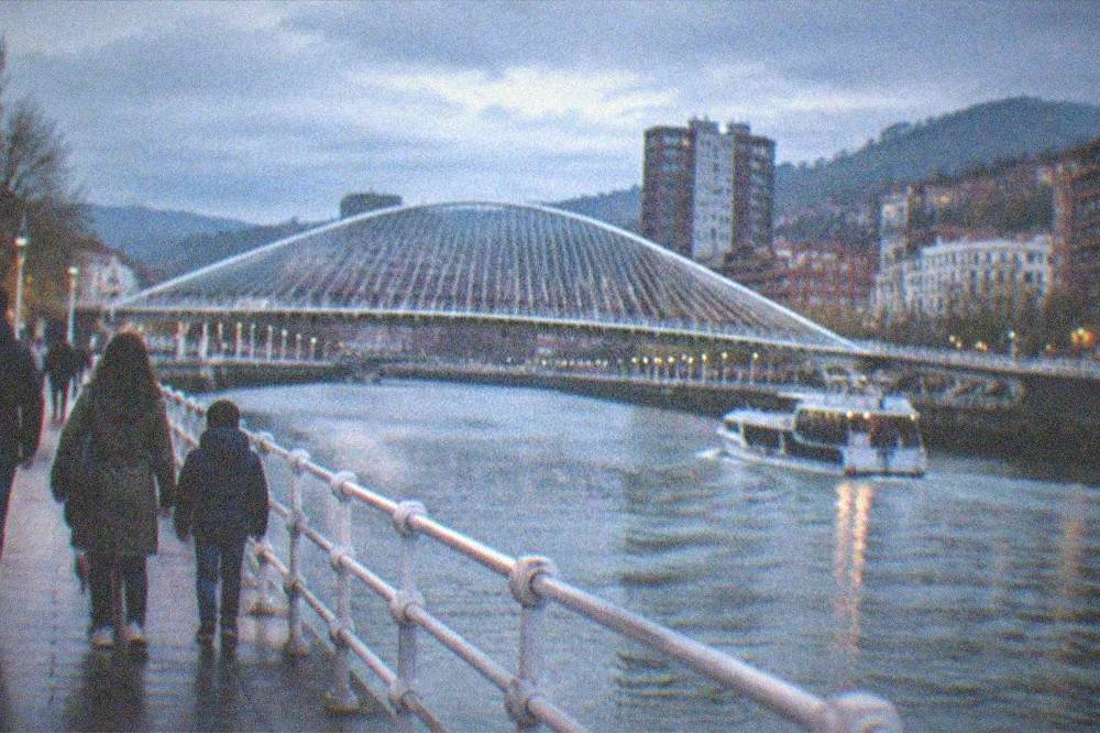 A family walking along the Nervion riverfront promenade in Bilbao with the Zubizuri bridge ahead