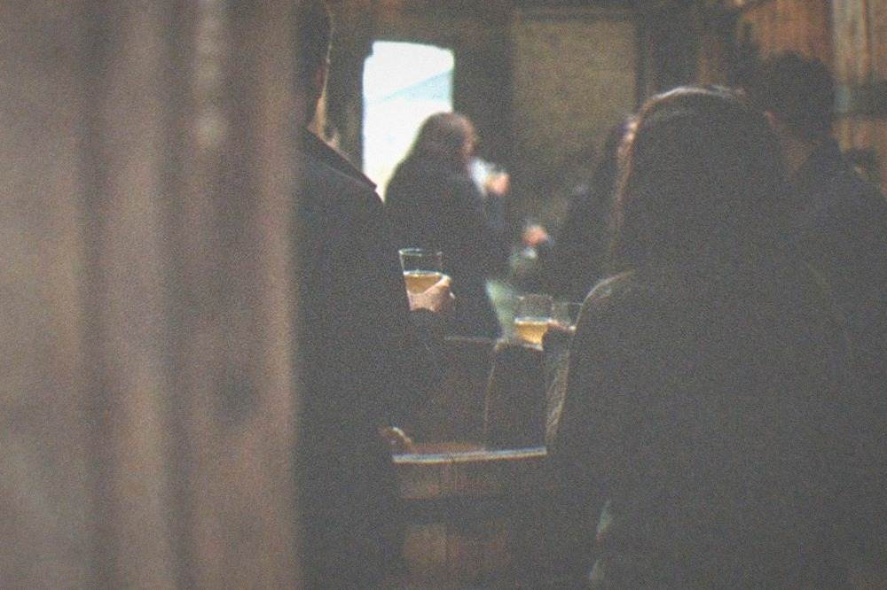 Guests standing around an open cider barrel holding glasses inside a dimly lit barrel room