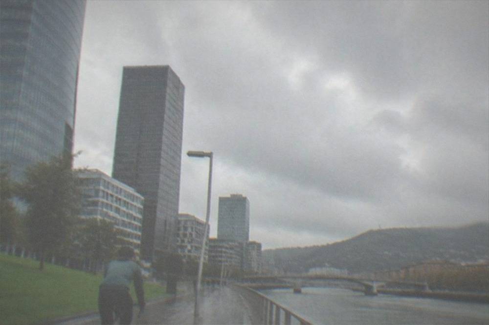 A road cyclist in a rain jacket riding along a damp riverside path with low clouds over green hills