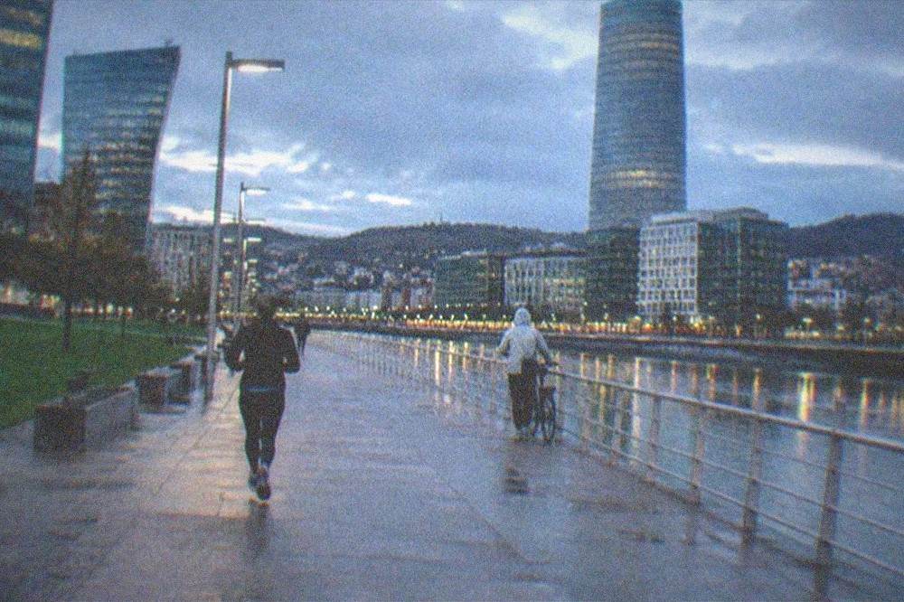A cyclist in a light rain jacket checking a map beside the estuary with modern buildings reflected in the water
