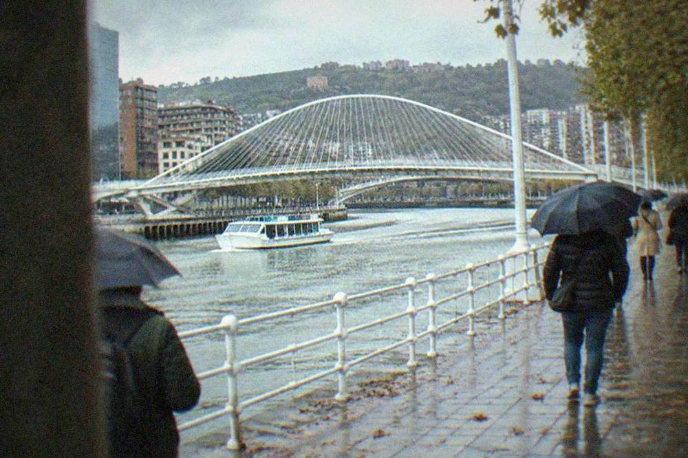 People walking with umbrellas past the Zubizuri bridge over the Nervion river in Bilbao on a rainy autumn day