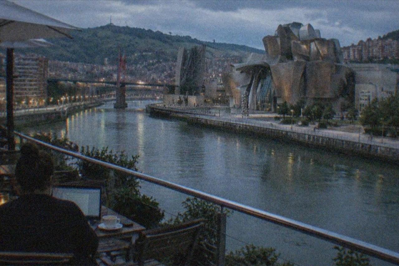A person with a laptop working at a cafe table near the Bilbao riverside with the Guggenheim Museum in the background
