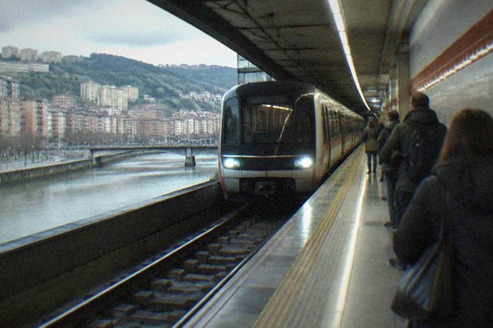 A Bilbao metro station platform with a train arriving and passengers waiting beside tiled walls