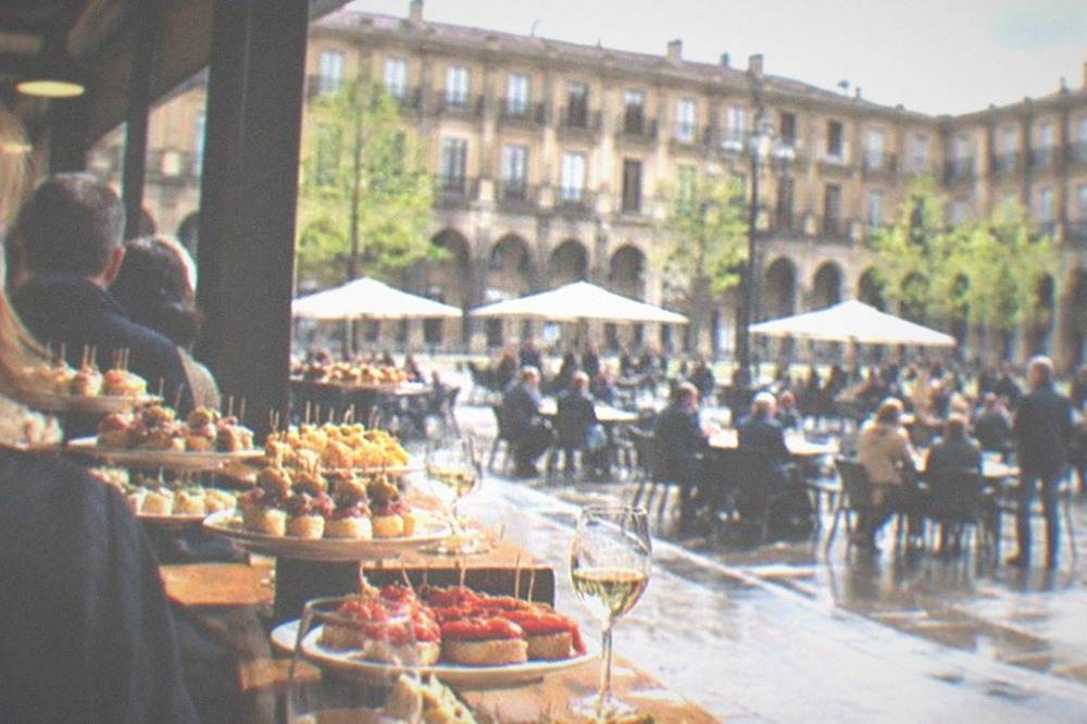 A crowded pintxos bar counter in Plaza Nueva with plates of small skewered bites and glasses of txakoli