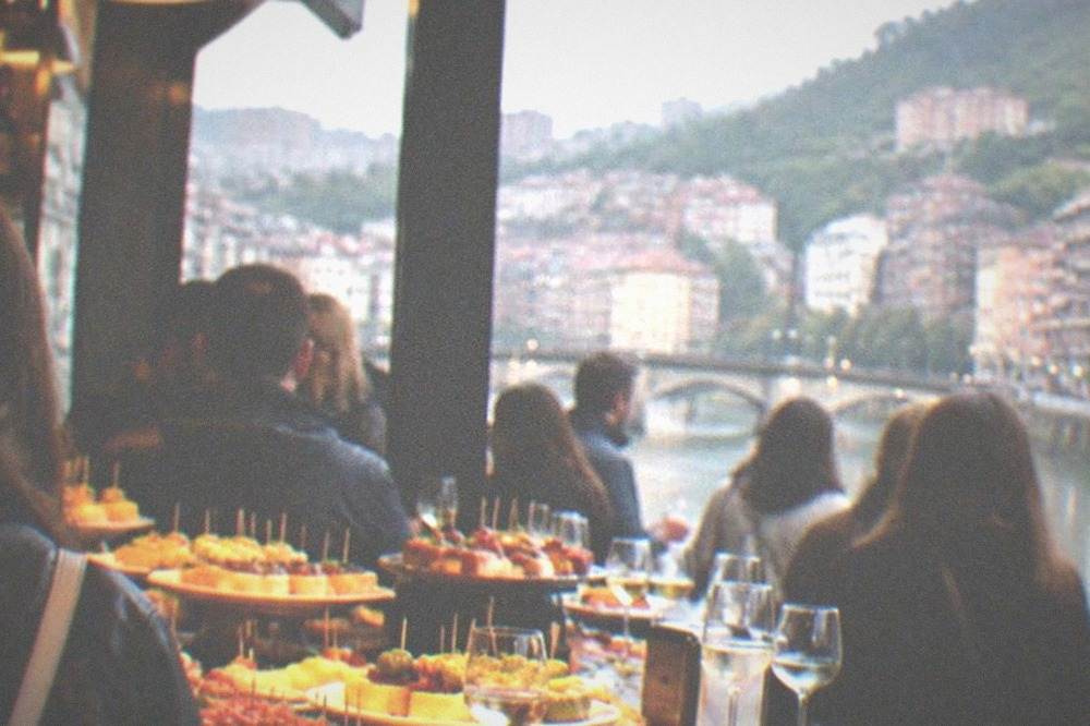 A crowded pintxo bar counter in Bilbao with plates of small skewered snacks and glasses of txakoli